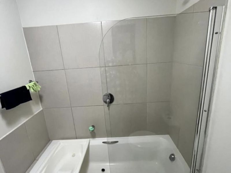 A clean bathroom featuring a built-in bathtub with a shower head, glass screen, and neutral wall tiling.