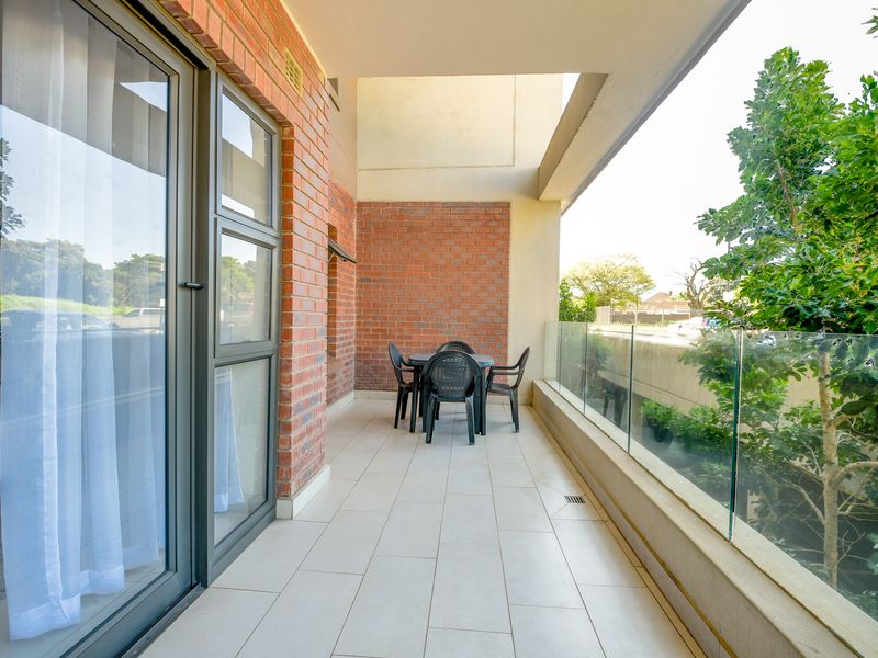 A spacious covered balcony featuring tiled flooring, an exposed brick wall, and a modern glass balustrade overlooking the surrounding greenery.