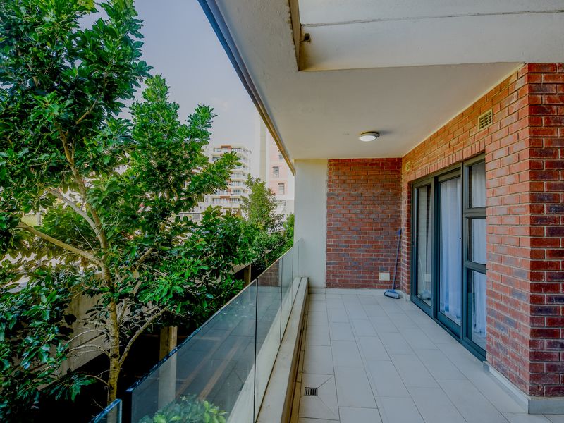 A spacious balcony featuring a modern glass balustrade, exposed brick wall, and tiled flooring, offering a pleasant outdoor area accessible via sliding doors.