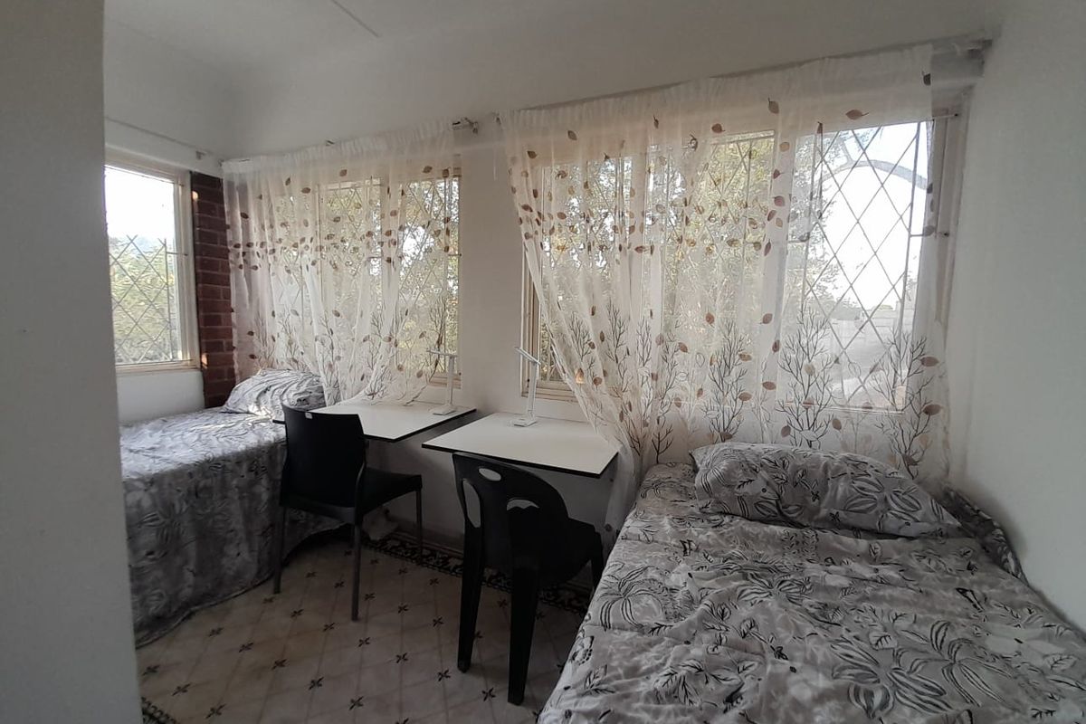 A functional bedroom featuring tiled flooring, exposed brick accents, and large windows fitted with security bars, providing ample natural light.