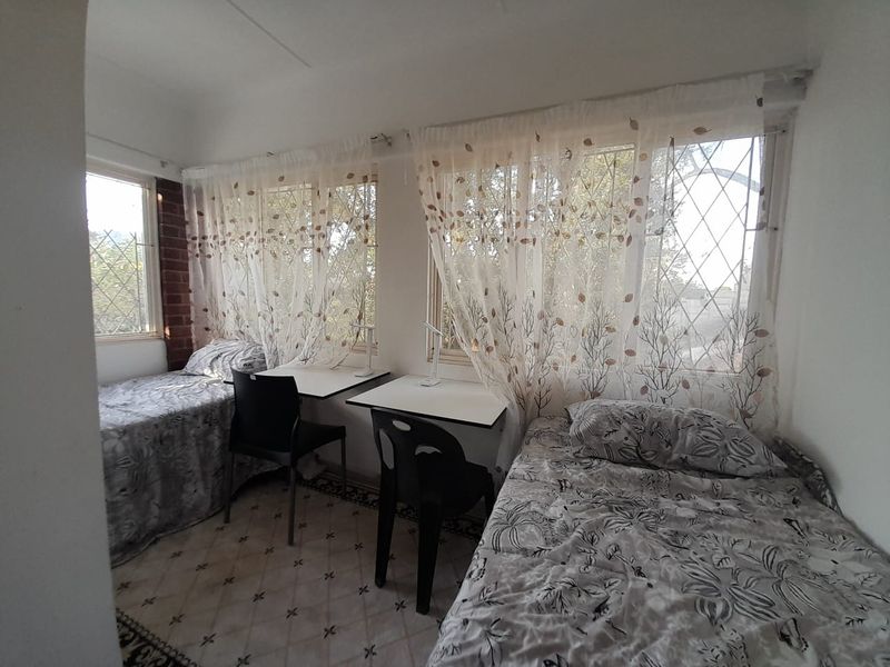A functional bedroom featuring tiled flooring, exposed brick accents, and large windows fitted with security bars, providing ample natural light.
