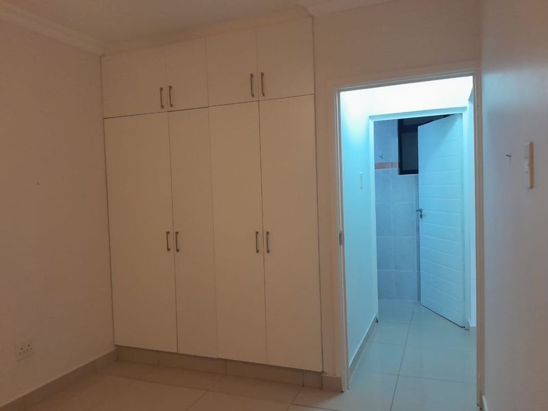 A clean, empty bedroom featuring white built-in cupboards, neutral tiled flooring, and direct access to an adjoining bathroom.