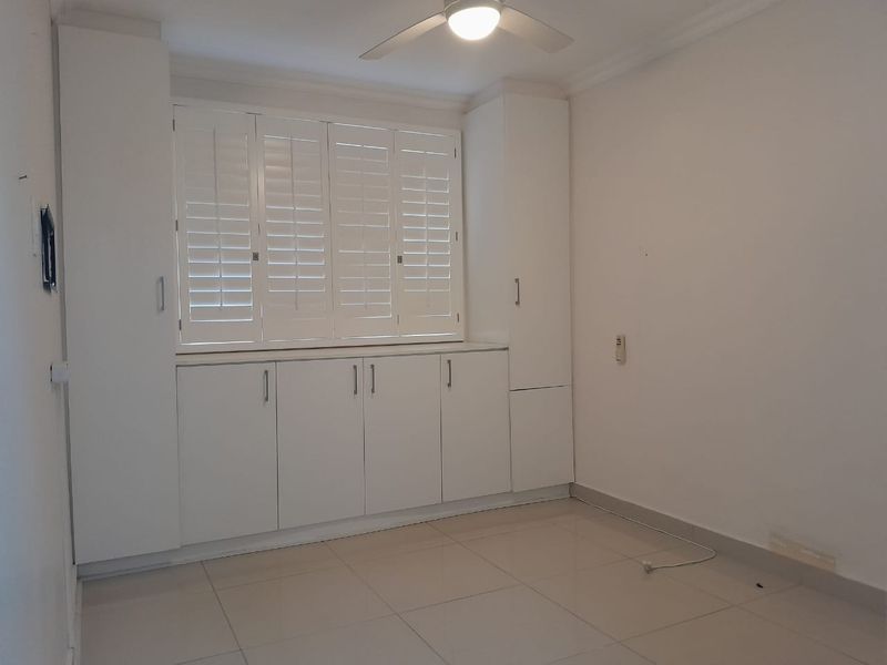 A bright, empty bedroom featuring neutral tiled flooring, extensive built-in cabinetry, and white plantation shutters for privacy and light control.