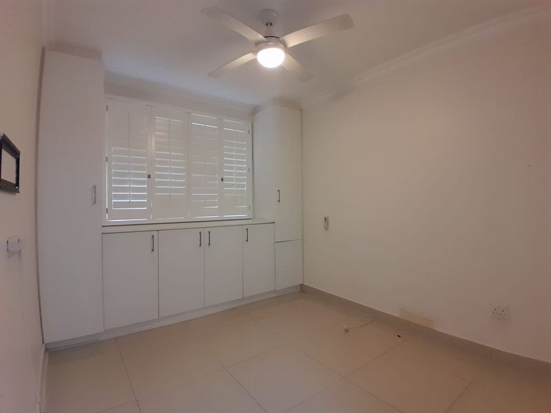 An empty bedroom featuring built-in cupboards, white plantation shutters, a ceiling fan, and neutral tiled flooring.