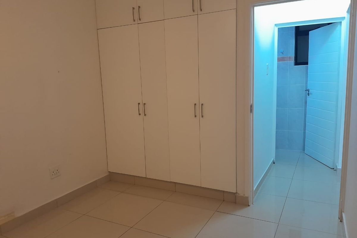 A clean, empty bedroom featuring white built-in cupboards and neutral tiled flooring, with an adjacent doorway leading to a tiled bathroom area.
