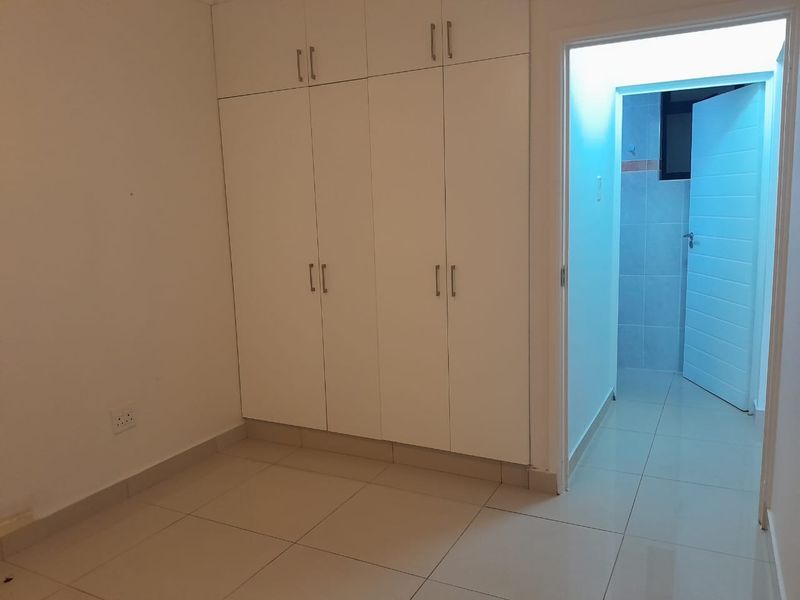 A clean, empty bedroom featuring white built-in cupboards and neutral tiled flooring, with an adjacent doorway leading to a tiled bathroom area.