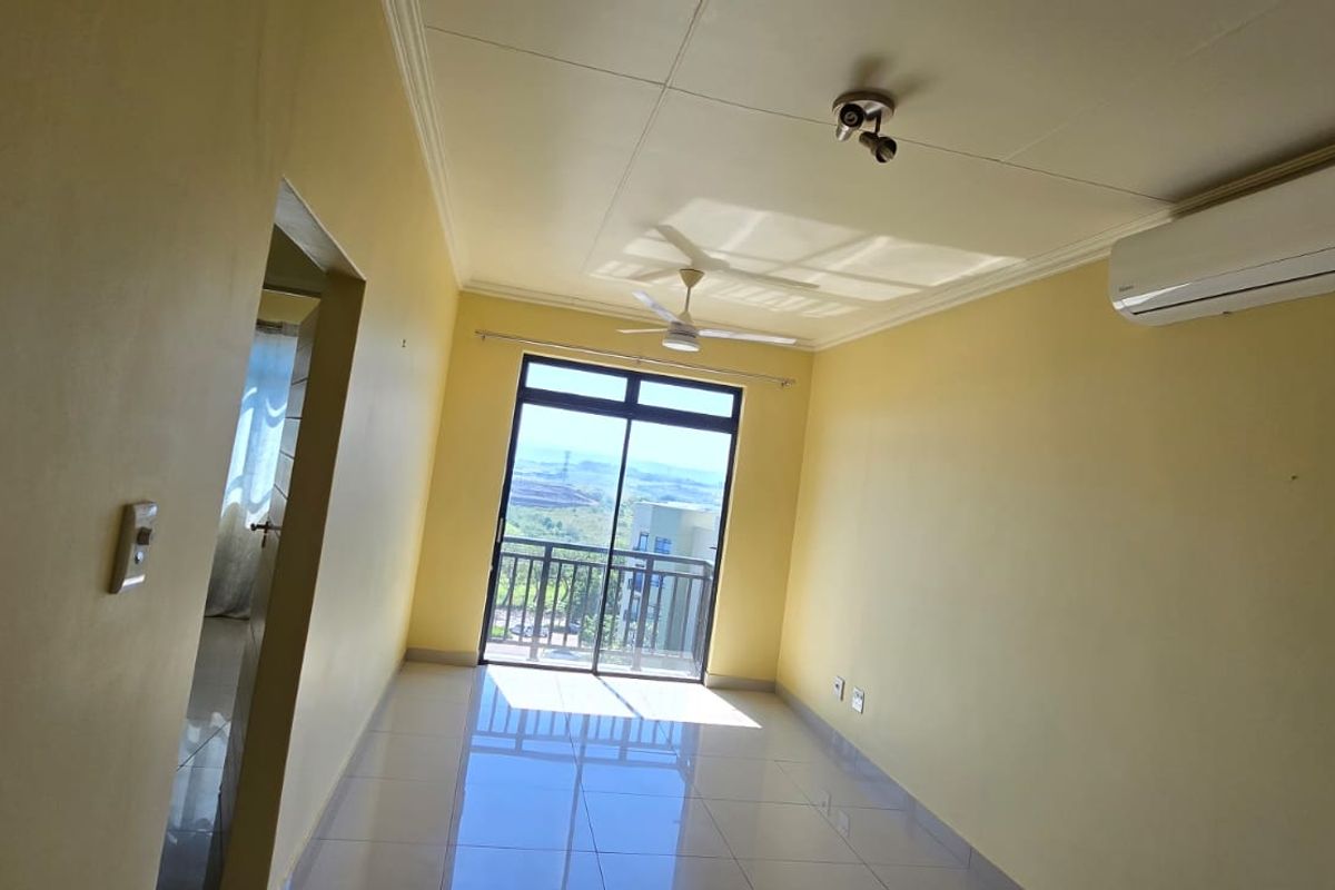 An empty, sunlit living room featuring tiled floors, a ceiling fan, a wall-mounted air conditioner, and access to a balcony with scenic views.