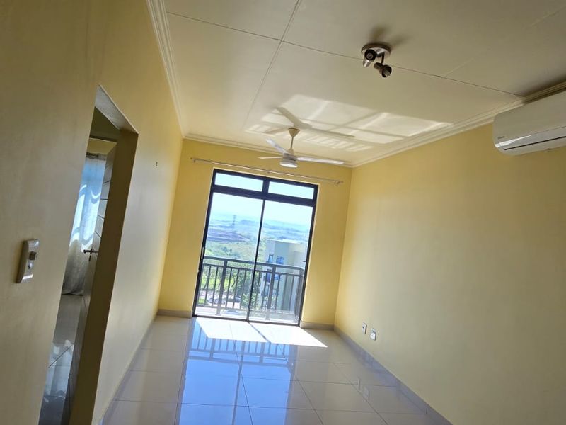 An empty, sunlit living room featuring tiled floors, a ceiling fan, a wall-mounted air conditioner, and access to a balcony with scenic views.