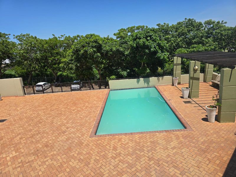 Outdoor pool area featuring a rectangular swimming pool set within a spacious brick-paved terrace, complemented by a pergola and mature surrounding trees.