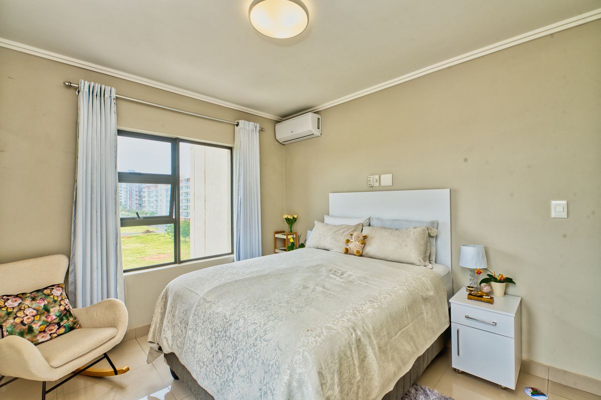 A well-lit bedroom featuring tiled flooring, a wall-mounted air conditioning unit, and large windows providing natural light and exterior views.