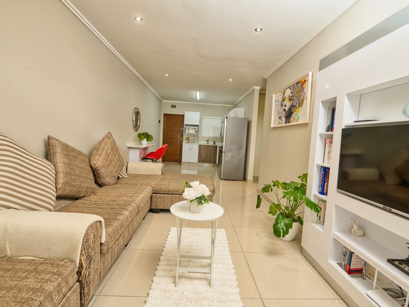 An open-plan living area featuring tiled flooring, recessed lighting, and a built-in media shelving unit, seamlessly connected to a functional kitchen space.