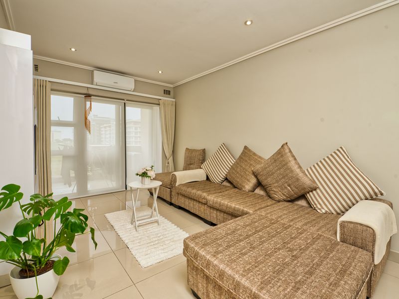 A bright and well-maintained living room featuring large format floor tiles, recessed lighting, and a wall-mounted air conditioning unit with access to a balcony.