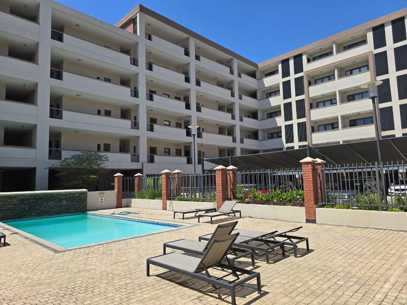 Communal pool area featuring a swimming pool with water feature, paved deck, and shade structure within a modern apartment complex.