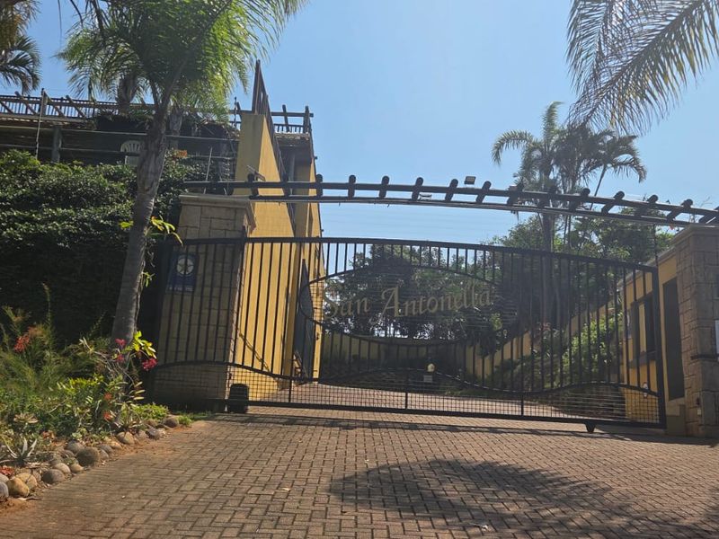 The entrance to San Antonella apartment complex featuring a secure electric gate, stone-clad pillars, and a paved driveway approach.