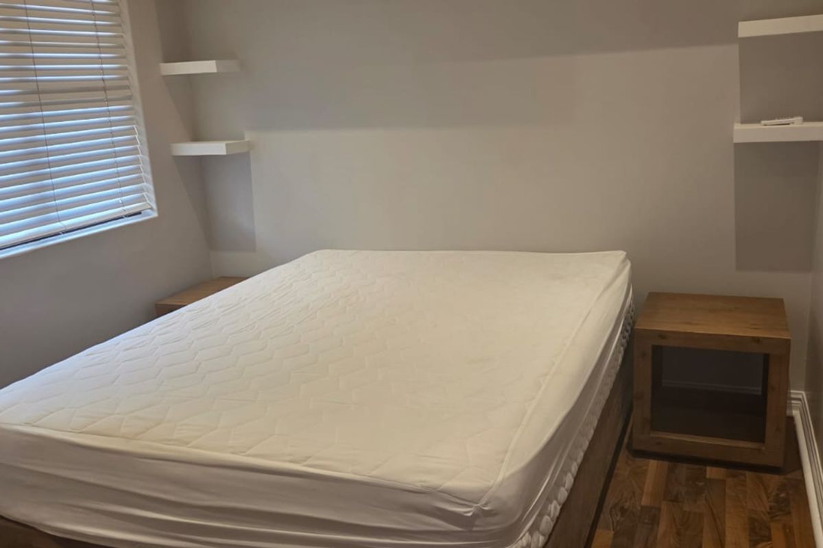 A compact bedroom featuring laminate flooring, built-in wardrobe storage, and wall-mounted floating shelves, finished with neutral tones and venetian blinds.