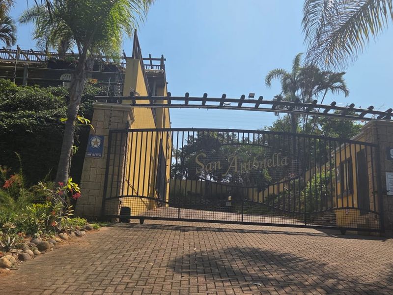 The entrance to San Antonella apartment complex featuring a secure gated access point with paved driveway and stone-clad perimeter walls.