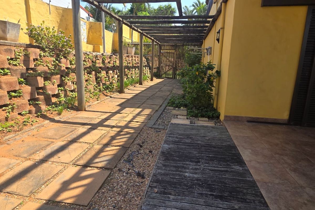 A rear exterior area featuring a paved walkway, timber pergola, and a tiered retaining wall with integrated garden beds.