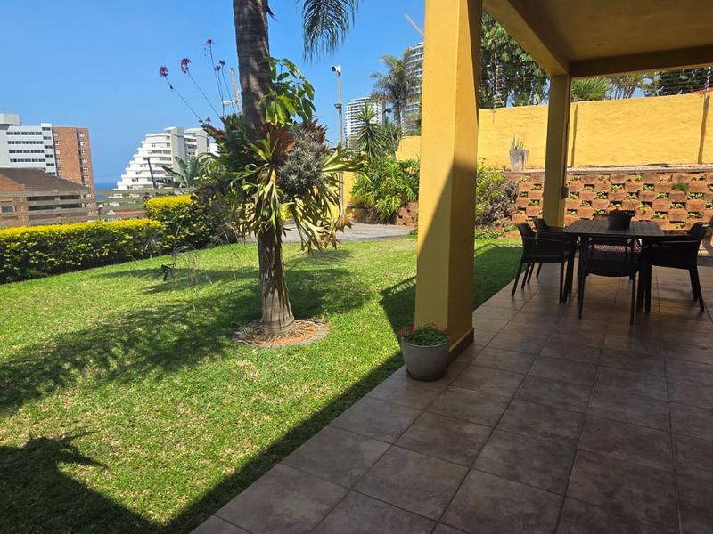 A covered patio area featuring tiled flooring, overlooking a private garden with a retaining wall and views of the surrounding neighborhood.
