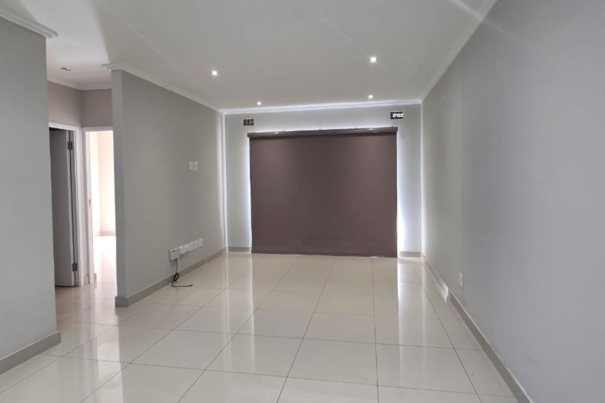 A bright, open-plan living area featuring neutral tiled flooring, recessed downlighting, and a modern roller blind.