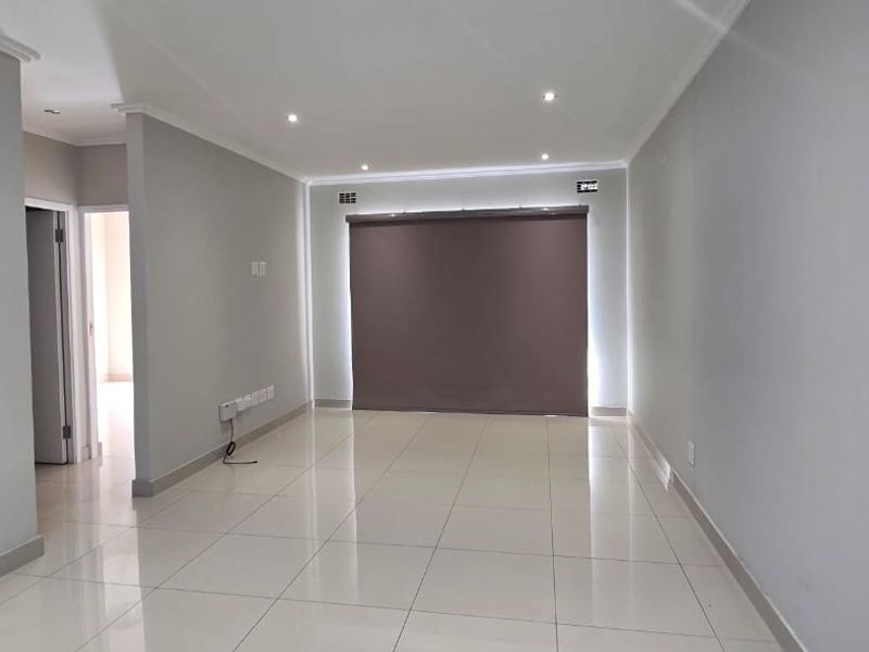 A bright, open-plan living area featuring neutral tiled flooring, recessed downlighting, and a modern roller blind.