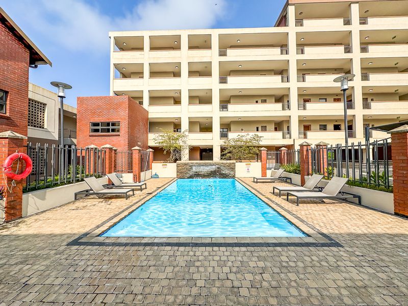 A well-maintained communal swimming pool area featuring a water feature, paved surround, and secure metal fencing within an apartment complex.