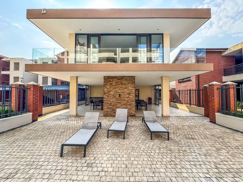 A modern outdoor patio area featuring a stone-clad feature wall, paved flooring, and a glass-balustraded balcony above, providing a contemporary communal space.