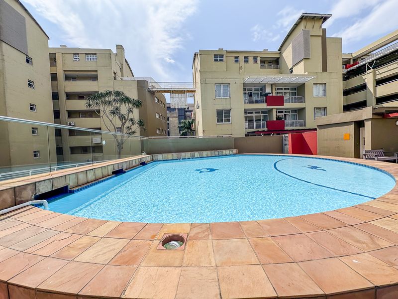 A communal swimming pool area featuring stone coping and a modern glass balustrade, set within an apartment complex courtyard.