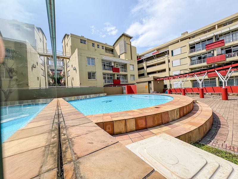 Communal swimming pool area featuring a tiled surround, paved courtyard, and pergola structure within an apartment complex setting.