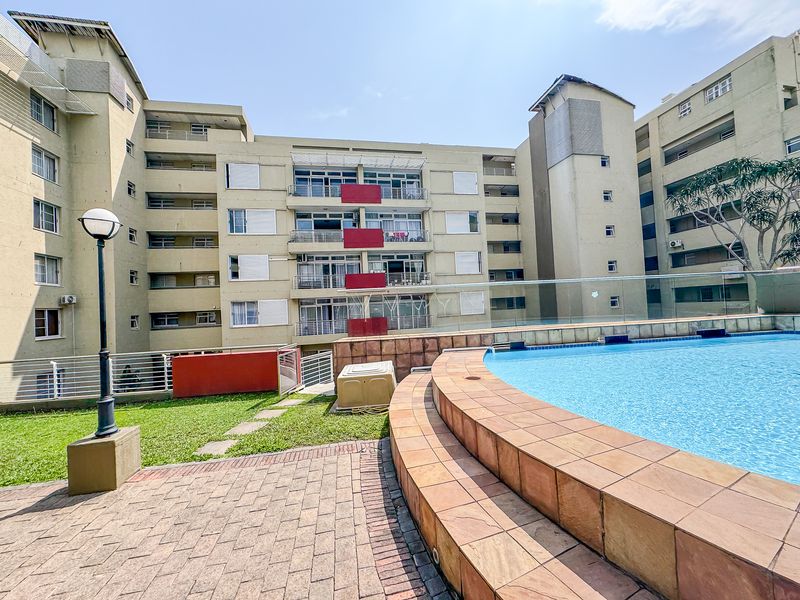 Communal pool area featuring a paved deck, glass balustrade, and landscaped lawn set against the apartment building facade.