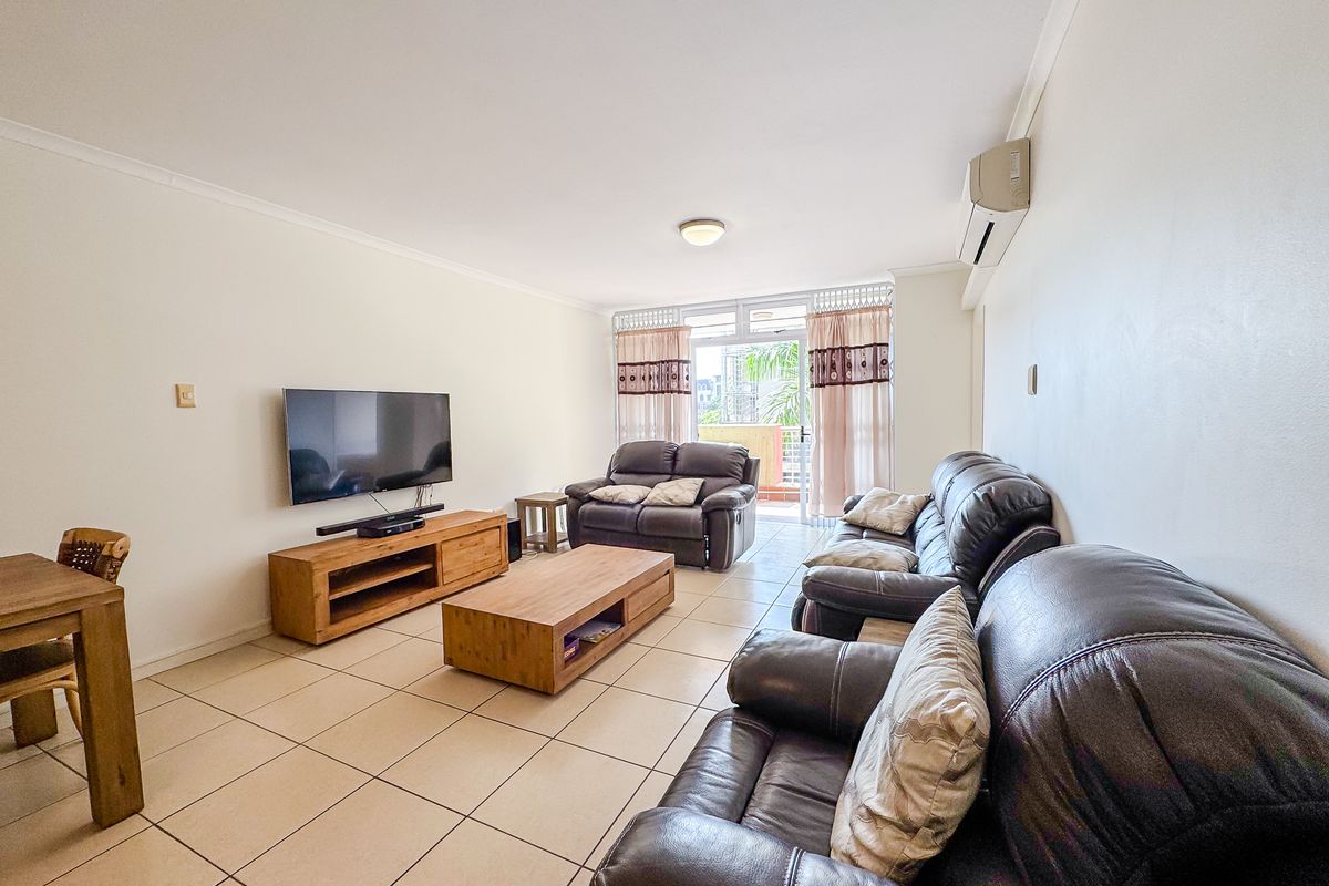 A spacious living room featuring neutral tiled flooring, a wall-mounted air conditioning unit, and direct access to an outdoor balcony through sliding glass doors.