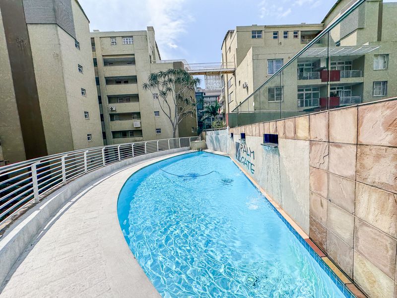 Communal swimming pool area featuring a water feature, stone wall cladding, and glass balustrades set between apartment building blocks.