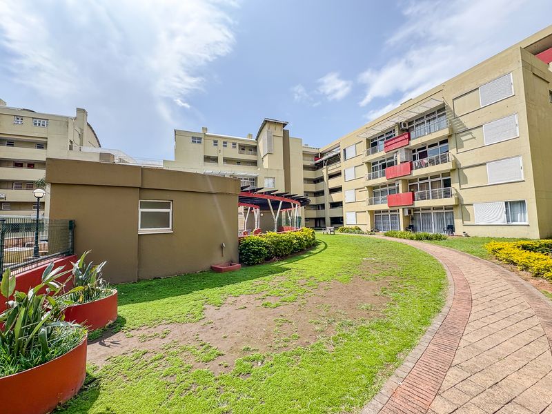 A view of the apartment complex exterior featuring a paved walkway, landscaped garden areas, a pergola, and multi-story residential units with balconies.