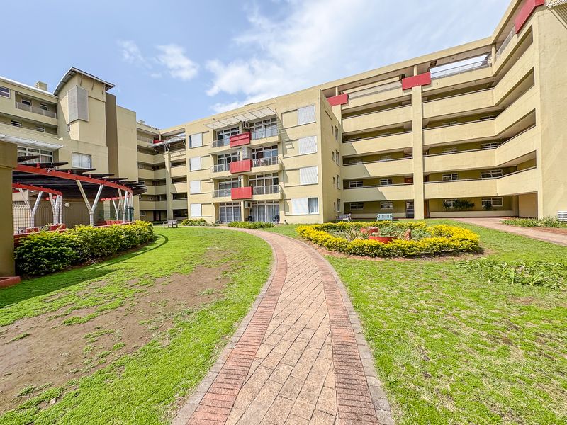 A view of the apartment complex exterior featuring a paved walkway, landscaped garden areas, and multi-level balconies.