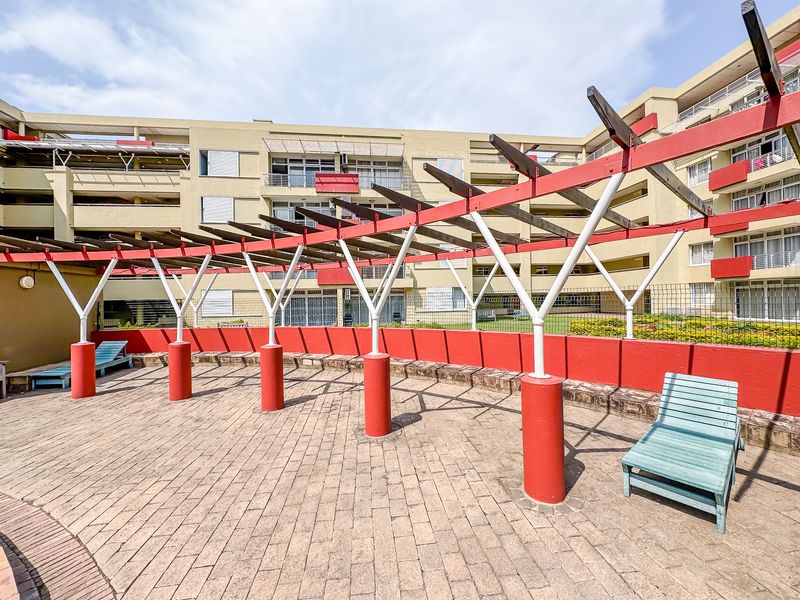 Outdoor pool area featuring a large decorative pergola, paved patio space, and a low retaining wall within an apartment complex setting.