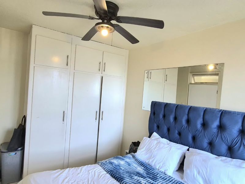 A bedroom featuring built-in cupboards, a ceiling fan, and a wall-mounted mirror, offering a functional layout with neutral finishes.