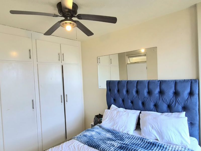 A functional bedroom featuring built-in cupboards, a ceiling fan, and a wall-mounted mirror, offering a practical layout with standard finishes.