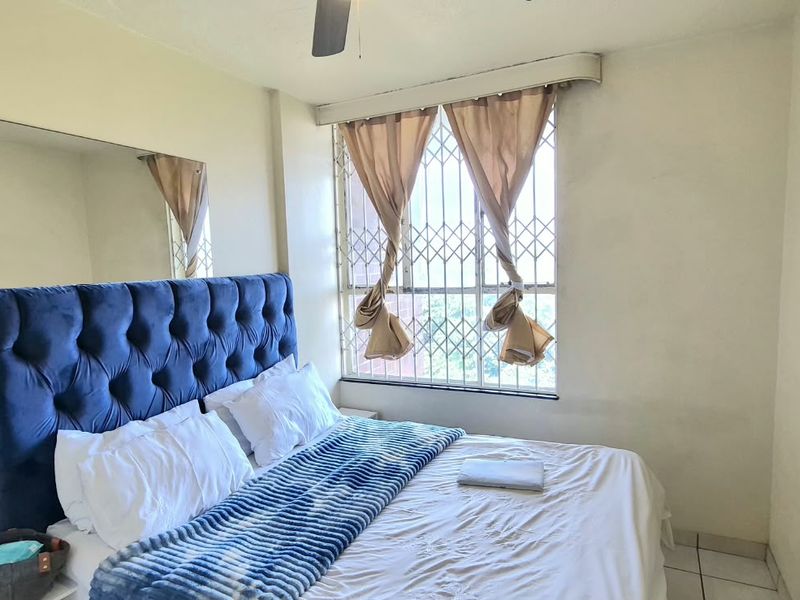 A bedroom featuring tiled flooring, a ceiling fan, and a window fitted with a security gate, offering a functional living space.