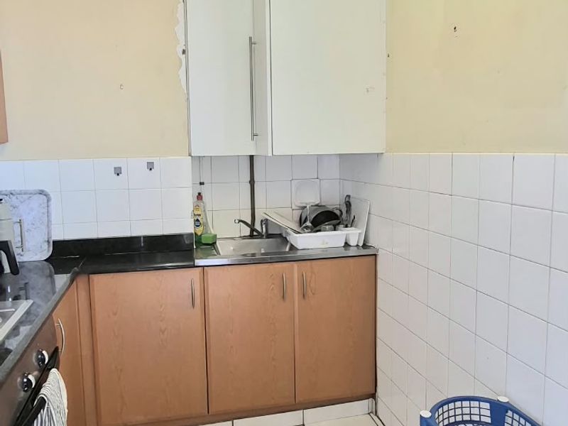 A functional kitchen featuring built-in cabinetry, a stainless steel sink, tiled backsplash, and tiled flooring, currently showing signs of wear and minor structural damage.