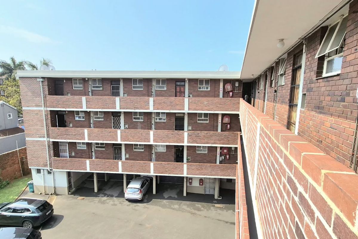 Exterior view of a multi-story brick apartment complex featuring open-air walkways, security gates, and ground-level covered parking facilities.