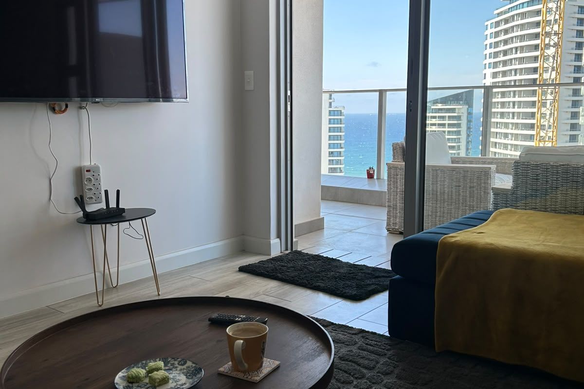 A bright living room featuring large sliding glass doors that open onto a balcony with scenic coastal and urban views.