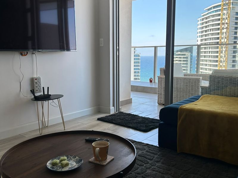 A bright living room featuring large sliding glass doors that open onto a balcony with scenic coastal and urban views.