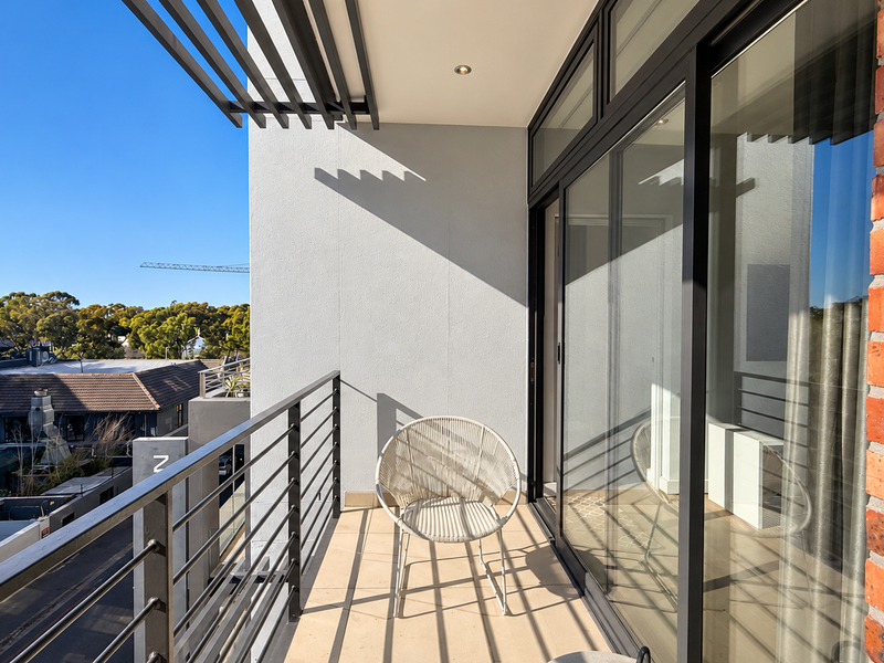 A modern apartment balcony featuring a metal balustrade, exposed brick accent wall, and sliding glass doors with overhead pergola detailing.