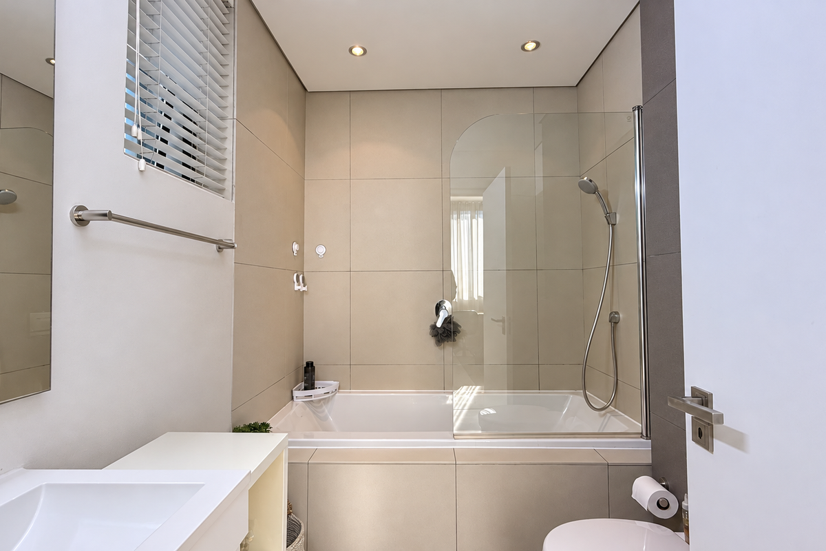 Modern bathroom featuring a built-in bathtub with a glass shower screen, neutral wall tiling, recessed lighting, and a wall-mounted vanity mirror.