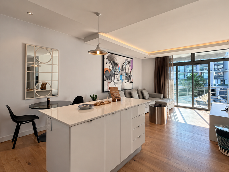 Modern open-plan living area featuring a central kitchen island with stone countertop, recessed lighting, and large sliding doors leading to a private balcony.