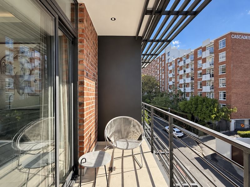 A modern apartment balcony featuring exposed brickwork, a sleek metal balustrade, and a slatted pergola, offering an urban view of the surrounding complex.