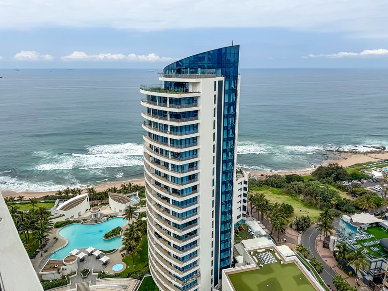 Aerial view of a modern high-rise apartment building featuring glass balconies, a swimming pool, and landscaped grounds overlooking the ocean.
