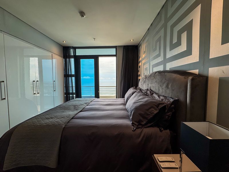 Modern bedroom featuring built-in cupboards, recessed lighting, and a sliding glass door providing access to a balcony with ocean views.