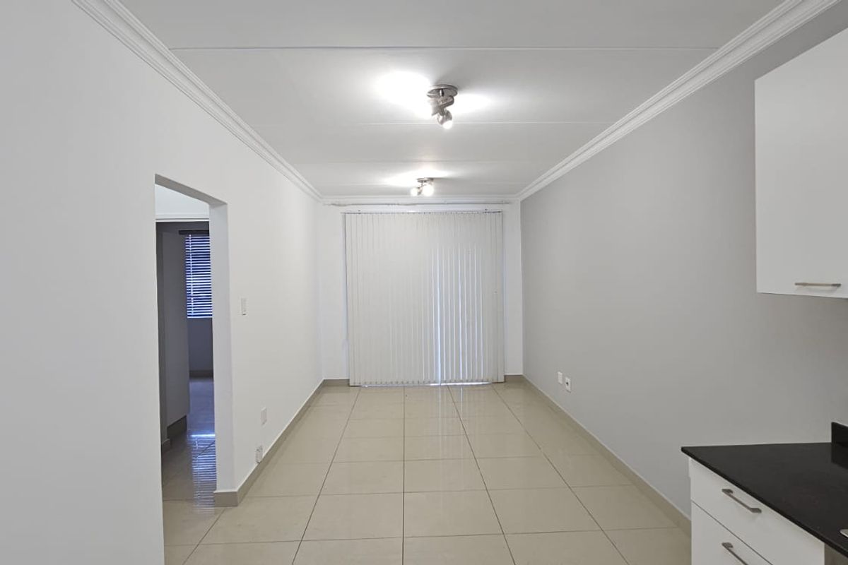 A bright, open-plan living and kitchen area featuring neutral tiled floors, modern cabinetry, and direct access to a balcony through vertical blinds.