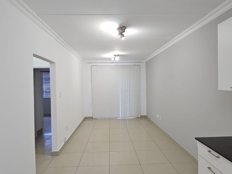 A bright, open-plan living and kitchen area featuring neutral tiled floors, modern cabinetry, and direct access to a balcony through vertical blinds.