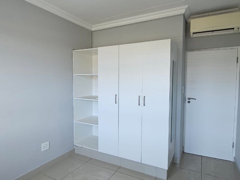 A clean, neutral bedroom featuring tiled flooring, a built-in wardrobe with open shelving, and a wall-mounted air conditioning unit.